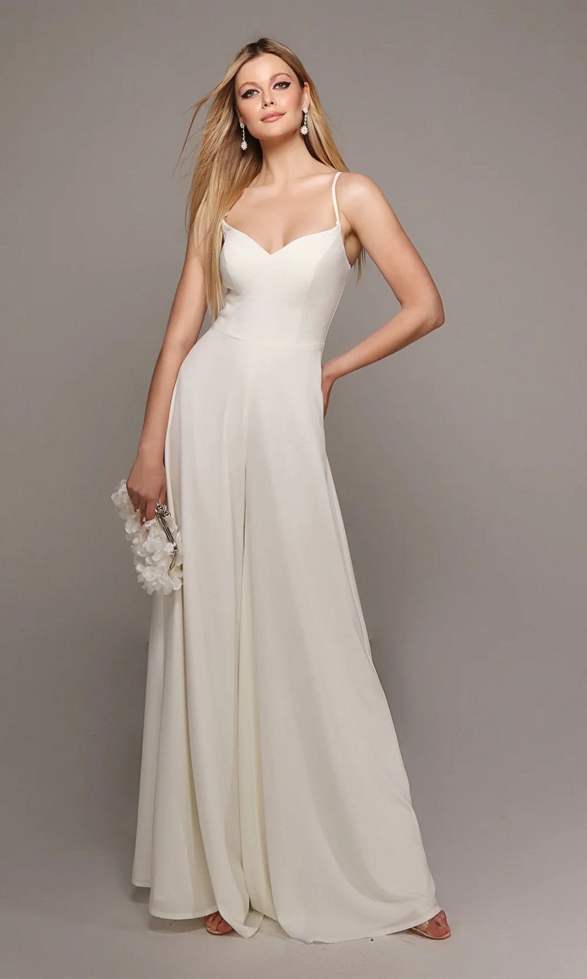 A woman poses elegantly against a plain gray background, wearing the Long White Jumpsuit: Alyce 70022 with wide-leg pants and holding a small bag.